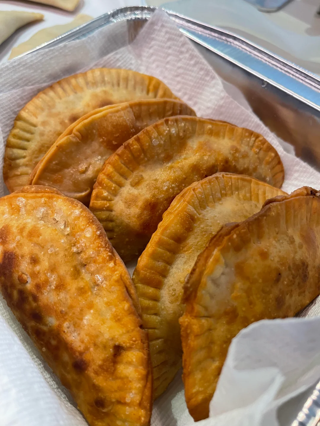 A close-up of several golden-brown empanadas arranged in a foil tray lined with white paper towels. The empanadas have crimped edges and are baked or fried, with a crispy texture.