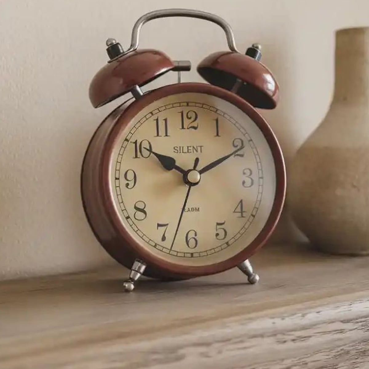 A vintage brown twin-bell alarm clock showing the time as 7:00 sits on a wooden surface next to a beige ceramic vase.
