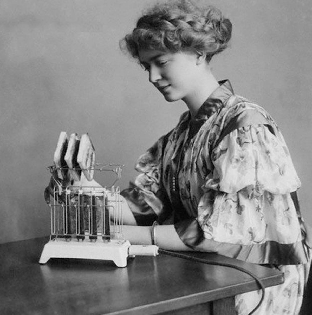A woman in early 20th-century clothing sits at a table, using a vintage toaster to toast slices of bread. She looks at the toaster attentively.