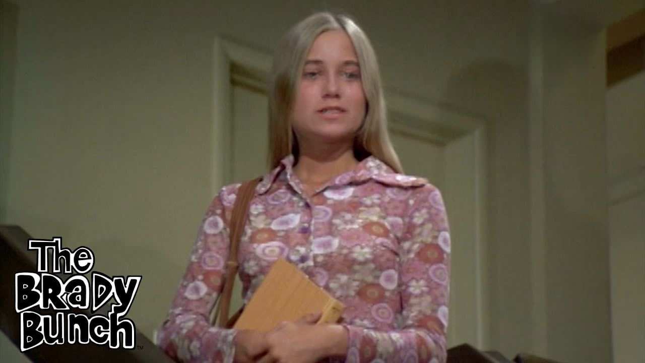 A young woman with long blonde hair, wearing a pink floral shirt, stands indoors holding a book. "The Brady Bunch" logo is in the lower left corner.