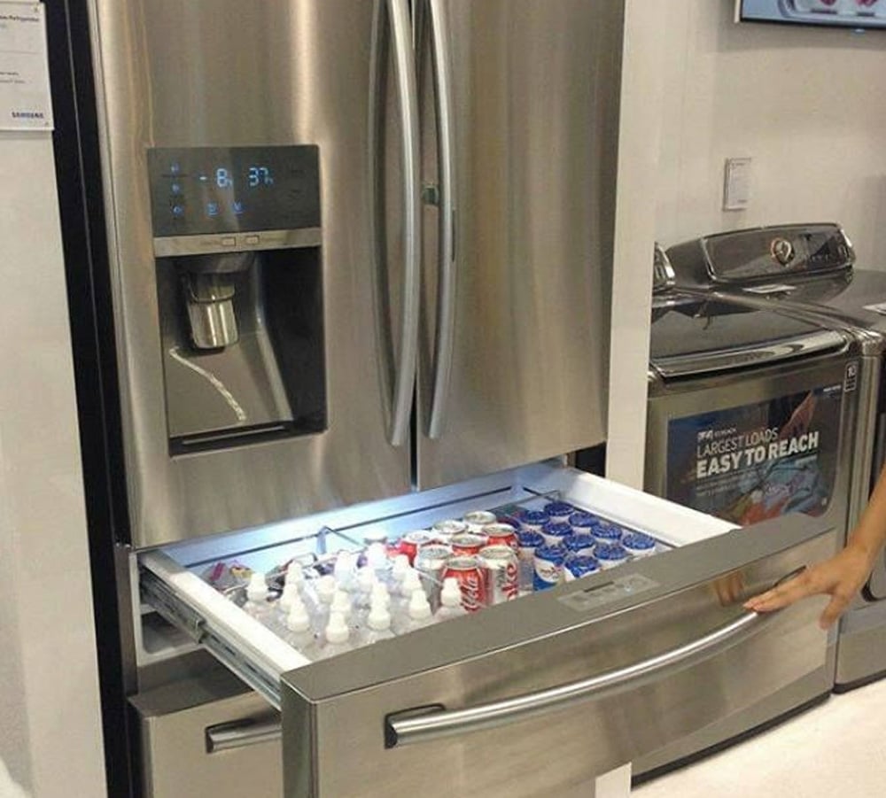 A refrigerator with a pull-out drawer open, showing cans of soda, small bottles, and ice packs neatly organized inside. A person’s hand is holding the drawer handle.