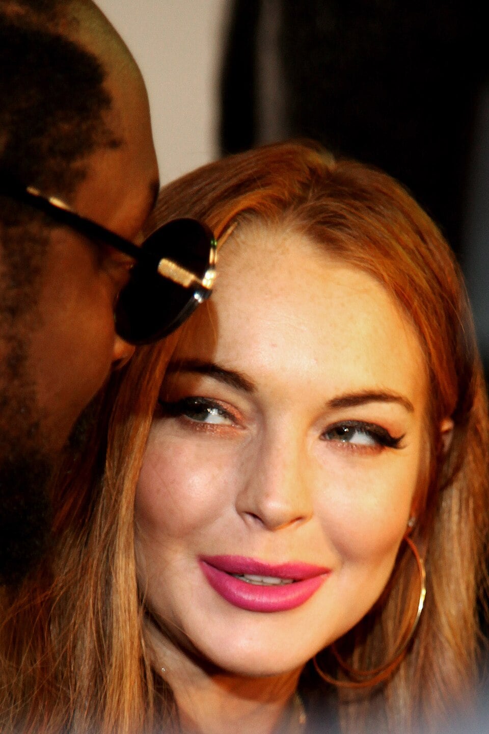 A woman with long red hair, bright pink lipstick, and gold hoop earrings smiles while looking at a man wearing dark sunglasses in the foreground.