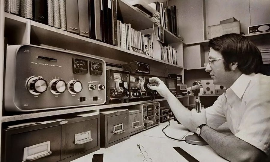 A man sits at a desk adjusting knobs on vintage radio equipment, surrounded by shelves filled with books, binders, and more electronics in a black and white setting.