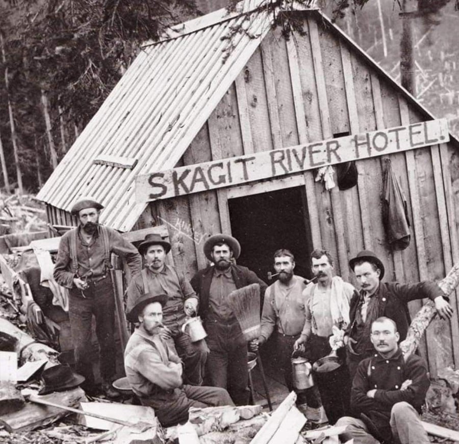 A group of men in work clothes and hats pose outside a small wooden cabin labeled “Skagit River Hotel,” surrounded by logs and trees in a rugged forest setting. Some hold mugs and tools, and most have facial hair.