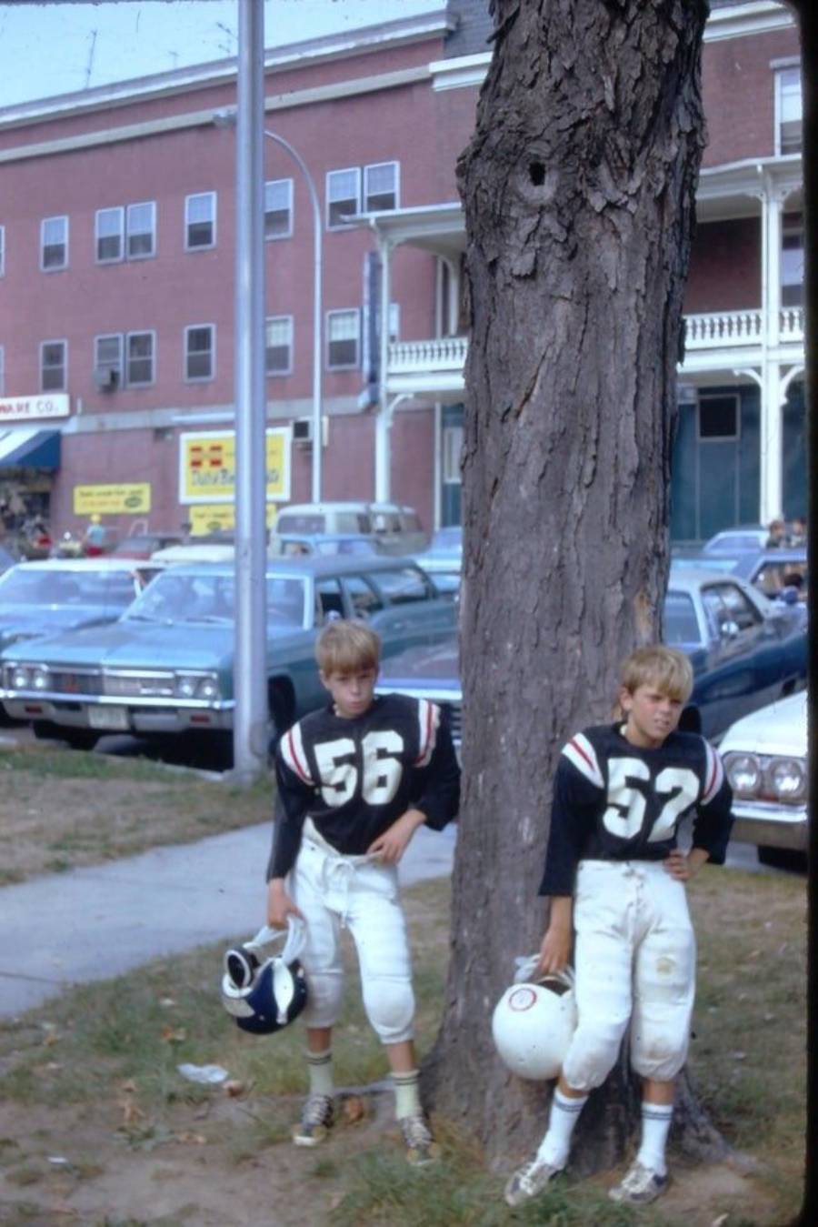 Two young boys in football uniforms, numbered 56 and 52, stand beside a large tree on a sidewalk. Both hold helmets, with cars and a brick building visible in the background.