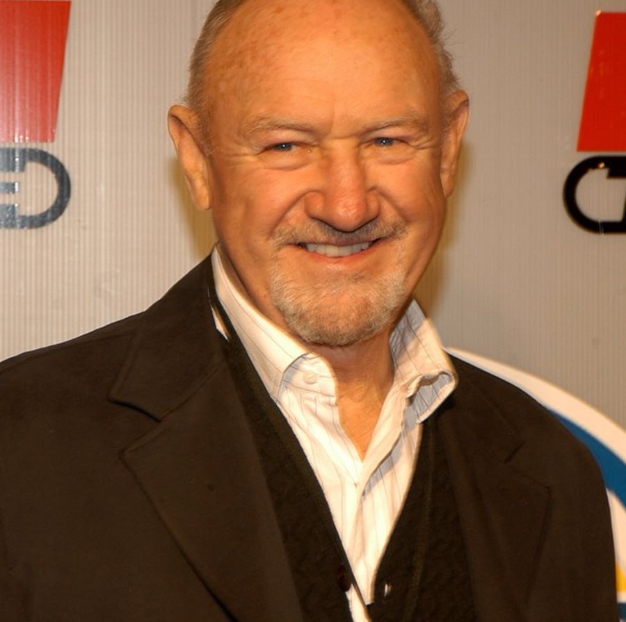 An older man with short gray hair and a trimmed beard is smiling. He wears a white collared shirt under a dark sweater and coat, standing in front of a backdrop with red and blue logos.