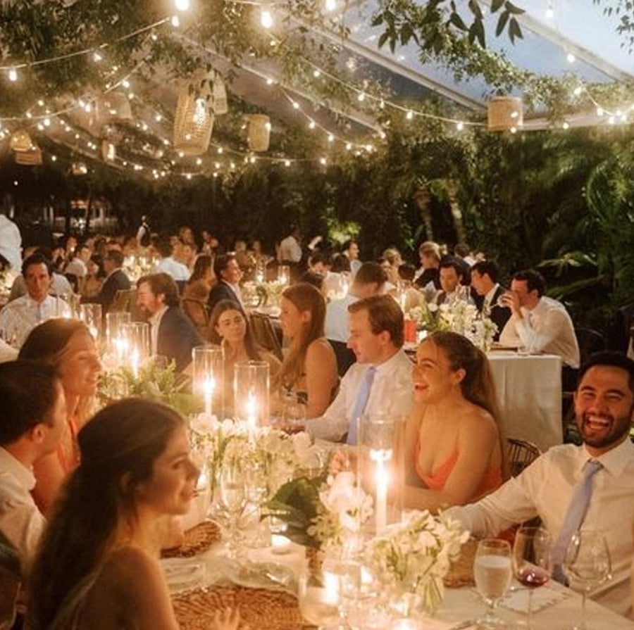 Guests sit at long tables decorated with candles and flowers at an elegant outdoor event under string lights, surrounded by greenery, creating a warm and festive evening atmosphere.