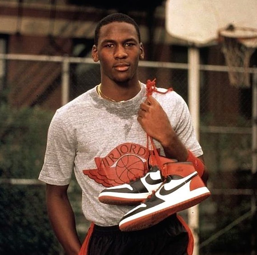 A young man in a gray "Air Jordan" t-shirt stands on an outdoor basketball court, holding a pair of red, white, and black Nike sneakers over his shoulder. A basketball hoop and fence are visible in the background.