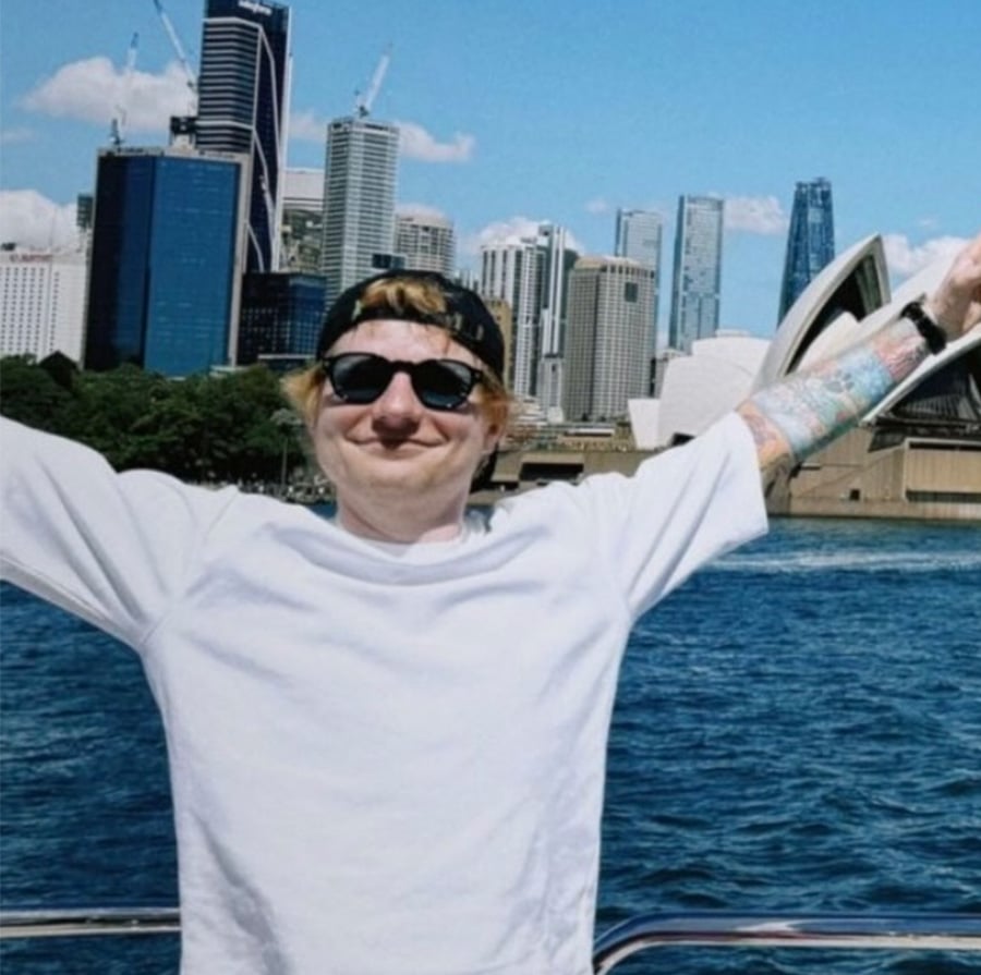 A person wearing sunglasses, a white shirt, and a backwards cap stands with arms raised in front of the Sydney Opera House and city skyline by the water on a sunny day.