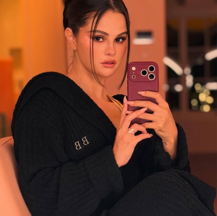 A woman with dark hair pulled back, wearing a black robe, sits indoors and takes a mirror selfie with a maroon phone, looking directly at the camera. Her robe has initials "B.B." embroidered on the chest.