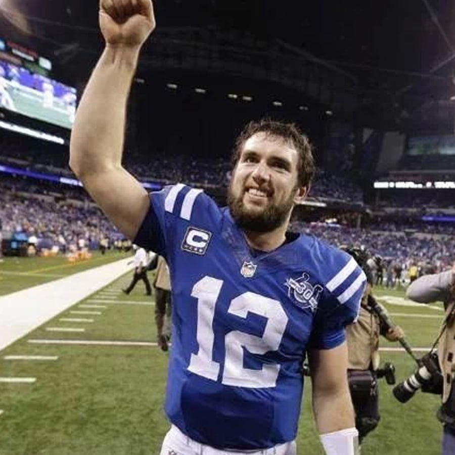 A football player wearing a blue jersey with the number 12 smiles and raises his right arm while standing on a football field, with a stadium and crowd in the background.