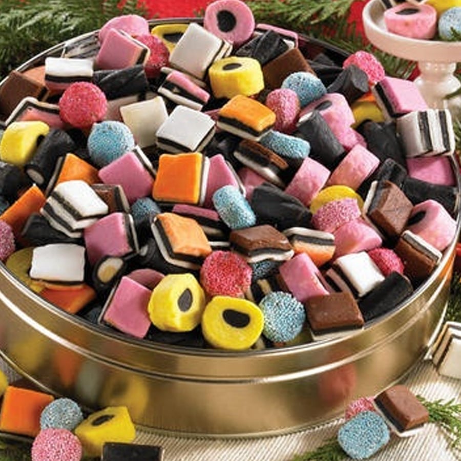 A round tin filled with colorful assorted licorice allsorts candies, including layered squares, round candies with sprinkles, and various shapes, set on a festive table with greenery accents.