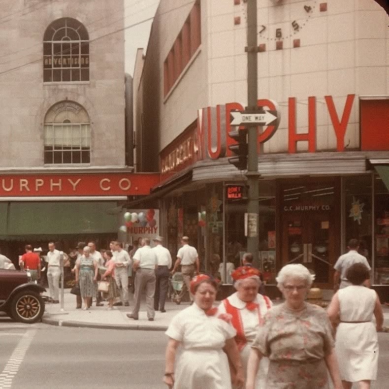 A busy 1950s city street corner with people walking, vintage cars, and a G.C. Murphy Co. store featuring a red and white sign and large display windows. The atmosphere is lively and bustling.