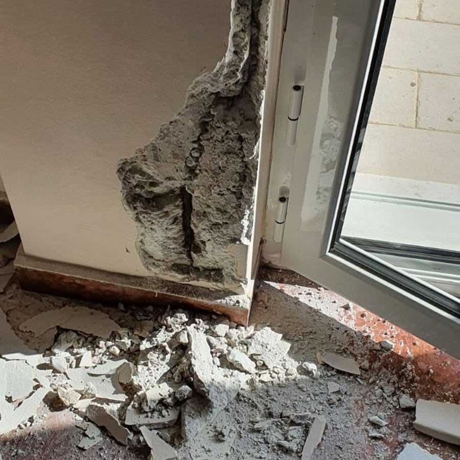 A wall near a door has a large, rough hole with plaster and debris scattered on the floor below, likely from recent damage or construction work. Sunlight shines through the open door.