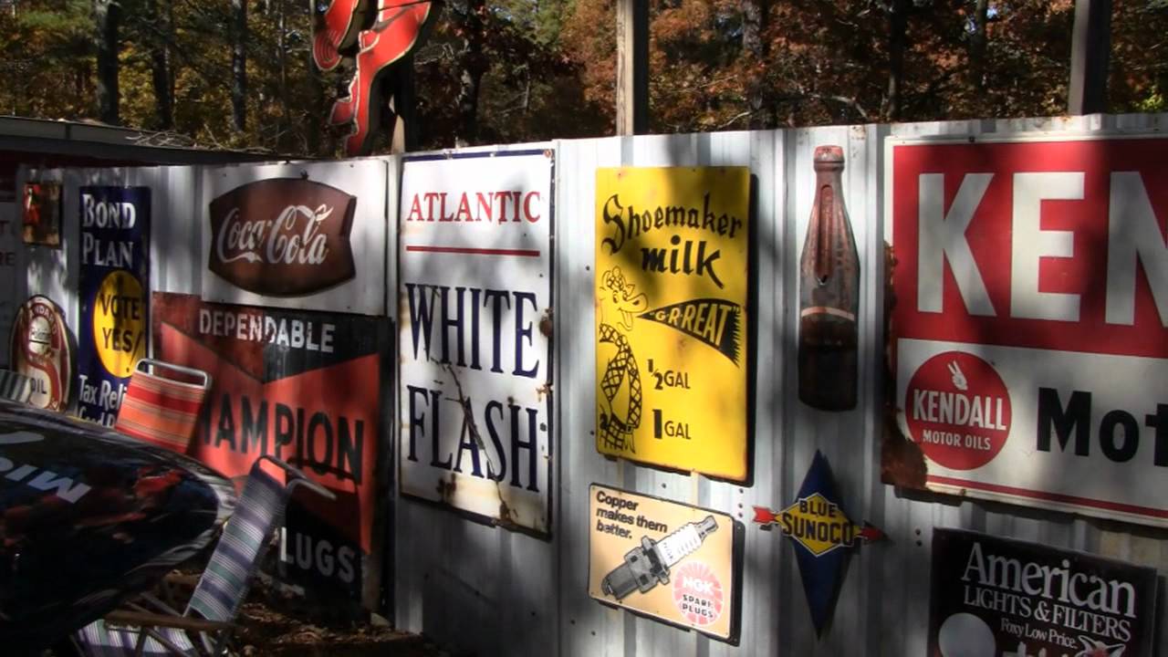 A collection of vintage metal advertising signs, including Coca-Cola, Shoemaker Milk, Kendall Motor Oils, and others, displayed on a wooden fence in an outdoor setting with sunlight and trees in the background.