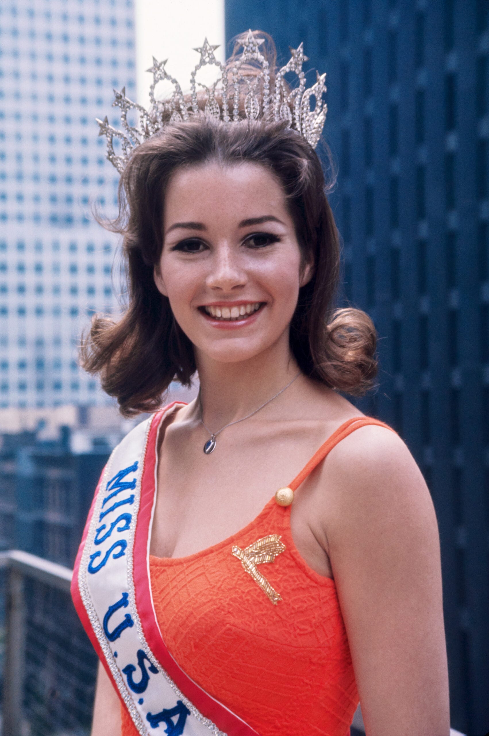 A smiling young woman wearing a jeweled crown and a "Miss U.S.A." sash stands outdoors in a city. She has shoulder-length brown hair and is dressed in an orange sleeveless top. Tall buildings are visible in the background.