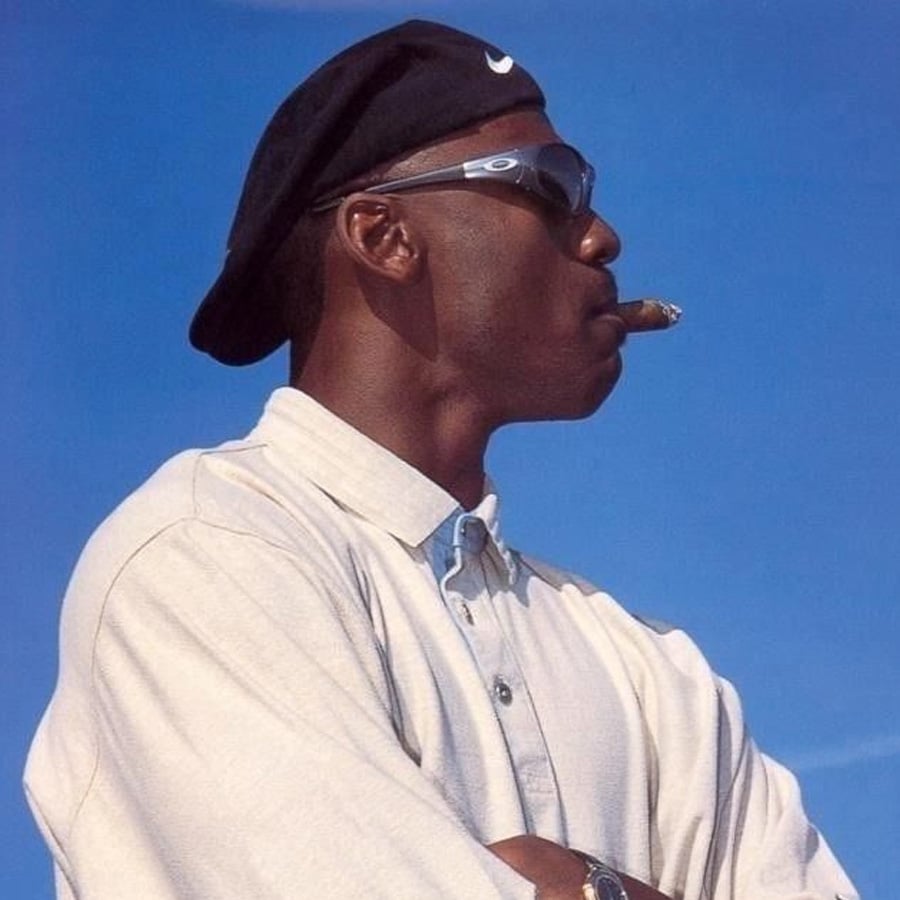 A man wearing sunglasses, a black cap, and a light-colored button-up shirt stands with his arms crossed, holding a cigar in his mouth, against a clear blue sky.