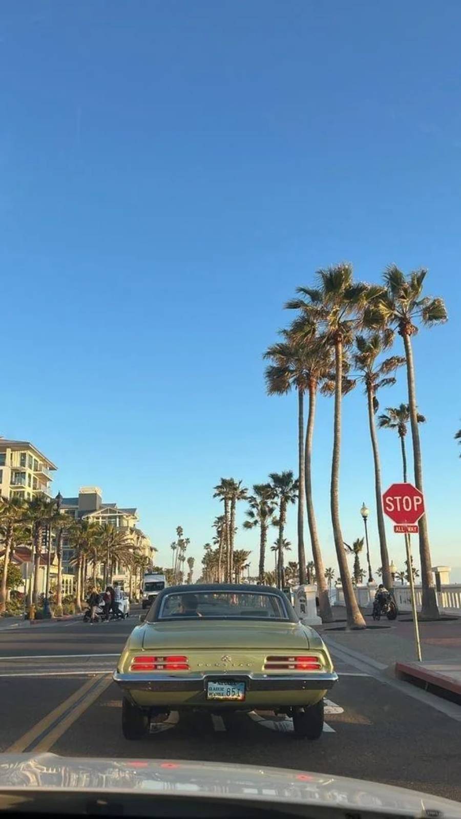 A vintage green car waits at a stop sign on a palm tree-lined street near the beach, with clear blue skies and people walking on the sidewalk.