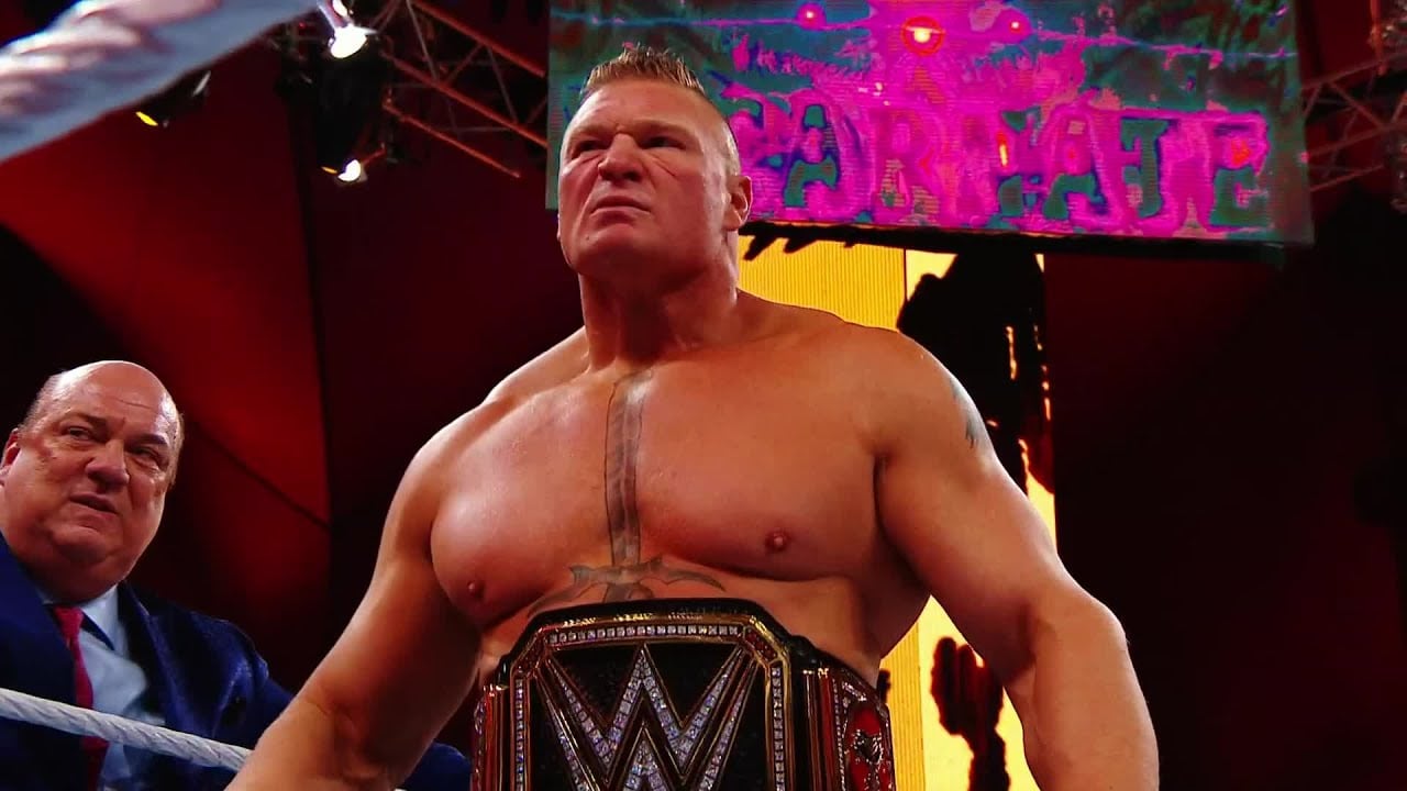 A muscular wrestler stands in a ring wearing a WWE championship belt, looking serious. Another man in a suit stands behind him. Bright lights and a colorful sign are visible in the background.