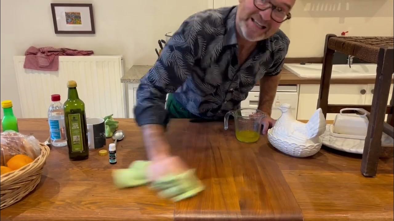 A man wearing glasses and a patterned shirt wipes a wooden kitchen counter with a green cloth. Various items, such as olive oil, a lemon juice bottle, and a basket of oranges, are on the counter.