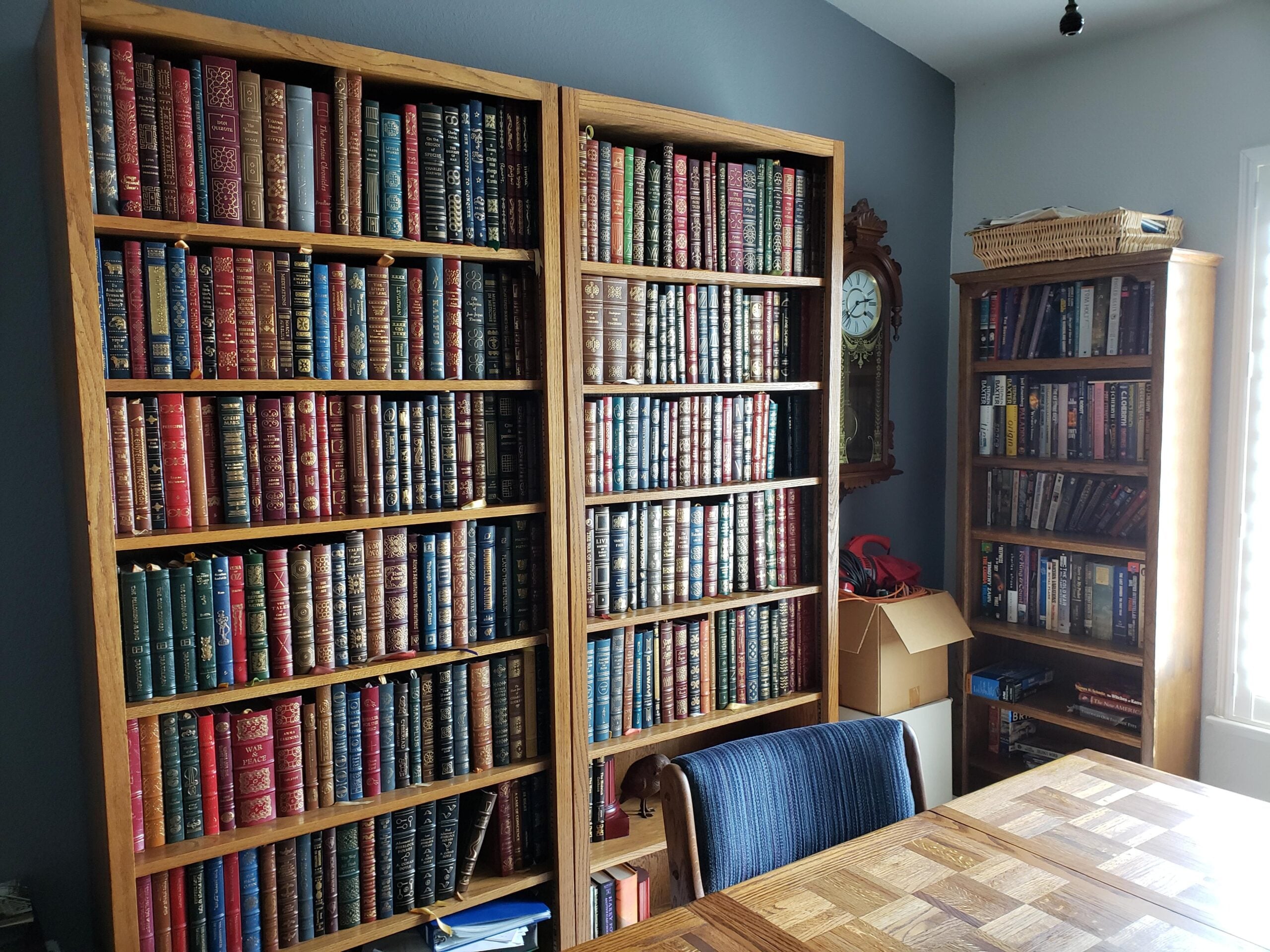 Three wooden bookshelves filled with colorful, mostly leather-bound books line a room with gray walls. A vintage clock hangs above a small table, and a checkered wood table and chair are in the foreground.