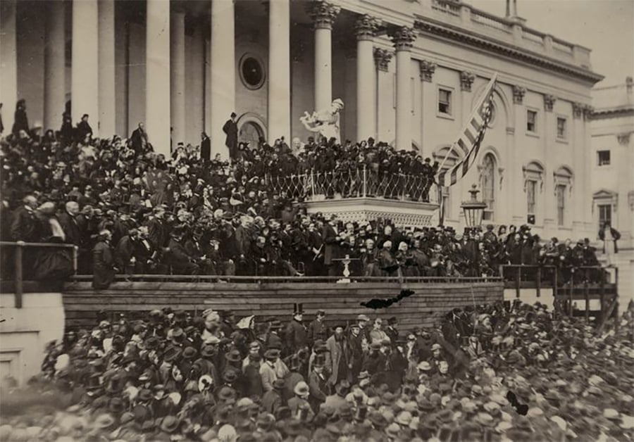 Large crowd gathered on the steps of a grand building with columns, watching a public event. An American flag is visible, and people are bundled in coats and hats. The scene appears historical and formal.