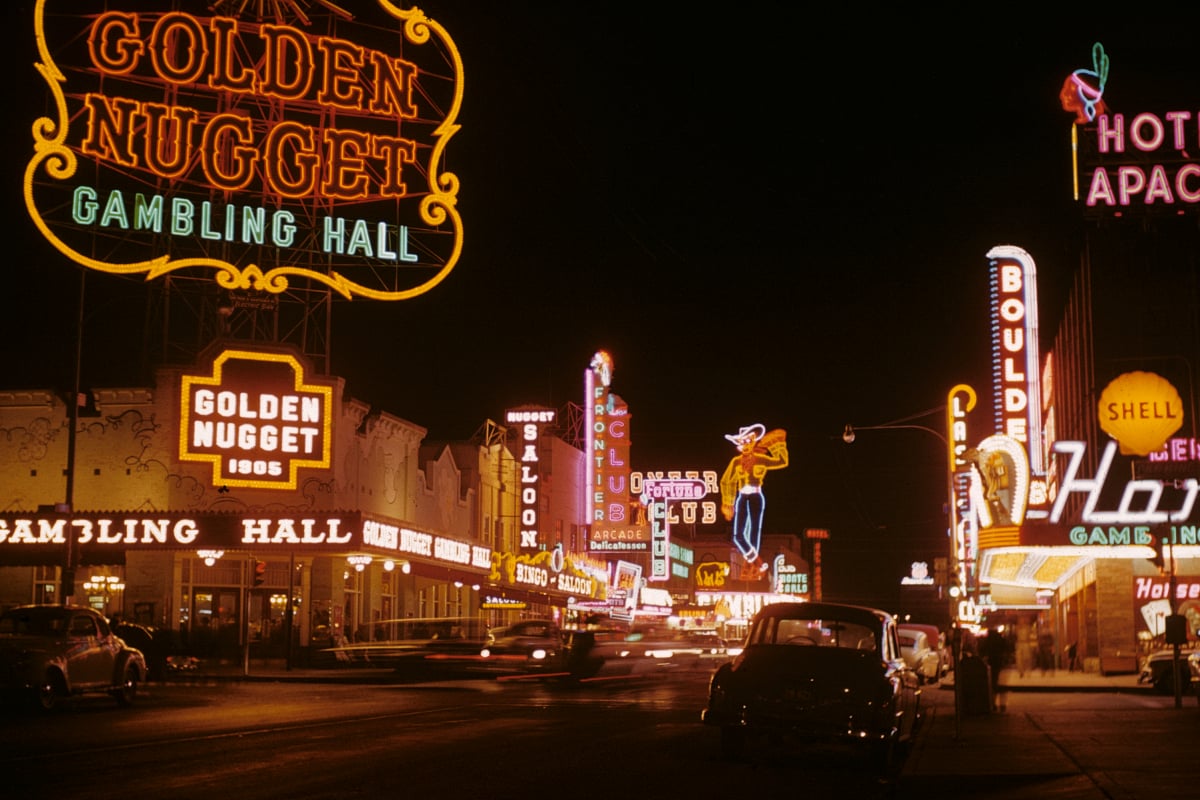 A vibrant night scene on a street lined with colorful neon signs, including "Golden Nugget Gambling Hall," "Hotel Apache," and "Boulder Club," with cars and people visible under the glowing lights.