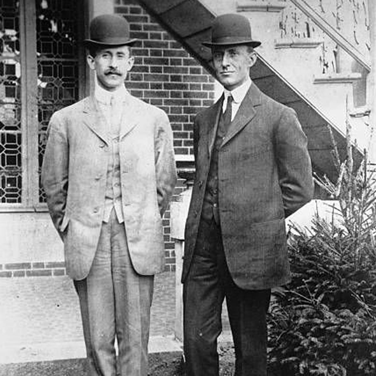 Two men in suits and bowler hats stand side by side outside a brick building with a window and staircase behind them. Both have mustaches and are posing formally for the photo.