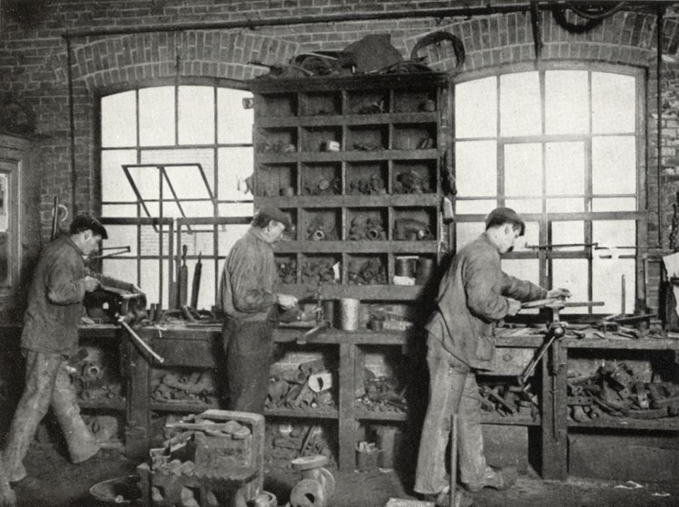 Three men work at a crowded workbench in a workshop filled with tools, pipes, and machine parts. Shelves and large windows are visible in the background, and the atmosphere appears industrious and busy.