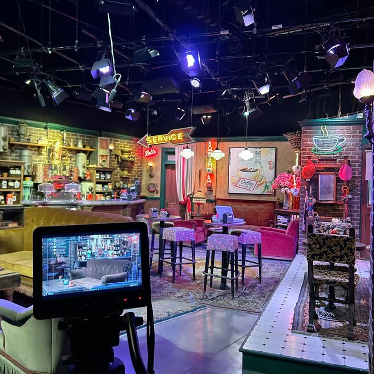 A brightly lit TV studio set designed as a cozy coffee shop, with vintage furniture, neon signs, tables with tablecloths, and shelves filled with mugs and decor. A camera monitor displays the set from a different angle.