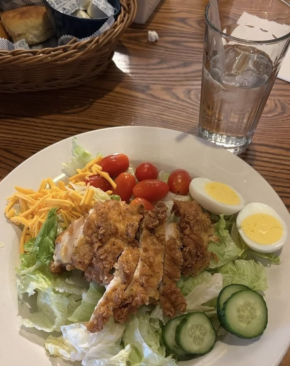 A plate with fried chicken strips on lettuce, sliced cucumber, cherry tomatoes, shredded cheese, and hard-boiled egg halves. A glass of ice water and a bread basket are on a wooden table in the background.