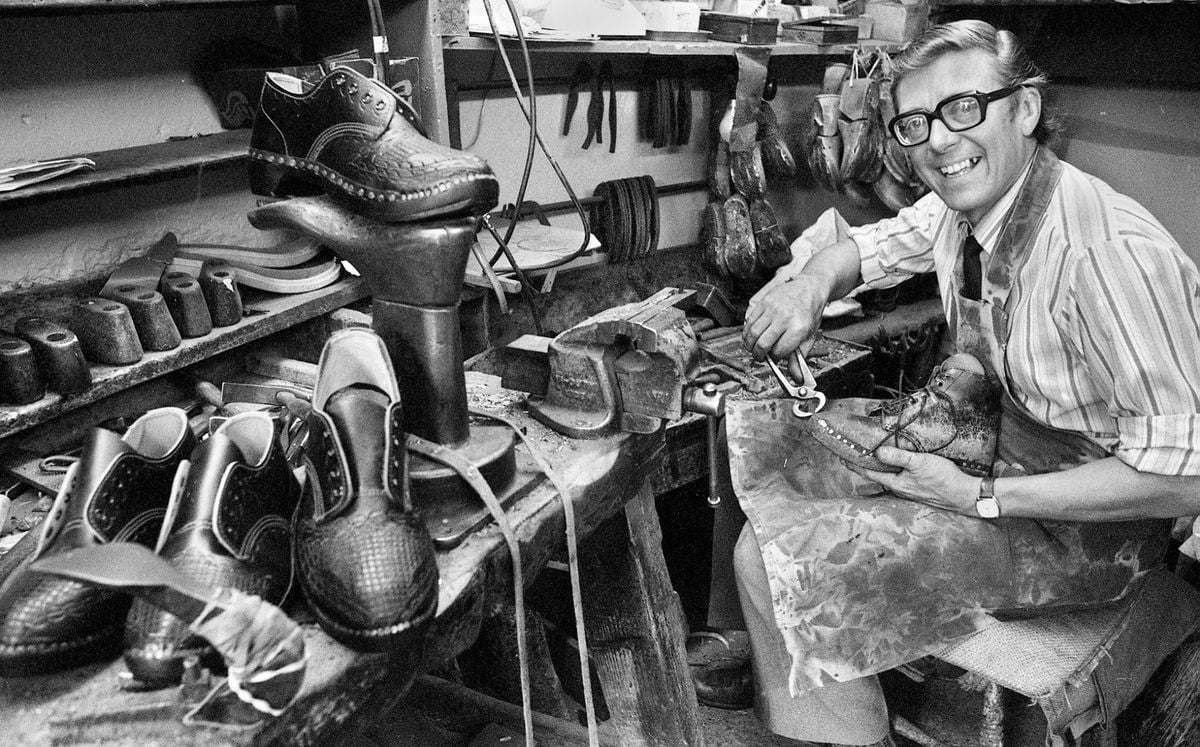 A smiling shoemaker sits at a cluttered workbench, crafting a shoe by hand. Tools, finished shoes, and shoe molds are scattered around the workspace. The setting appears to be a traditional shoe repair or crafting shop.