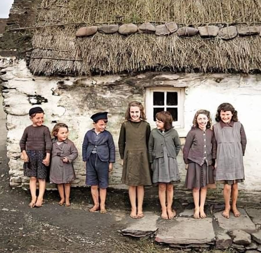 Seven barefoot children in old-fashioned, simple clothing stand in front of a stone cottage with a thatched roof. They are smiling and lined up on uneven ground, with a small window behind them.