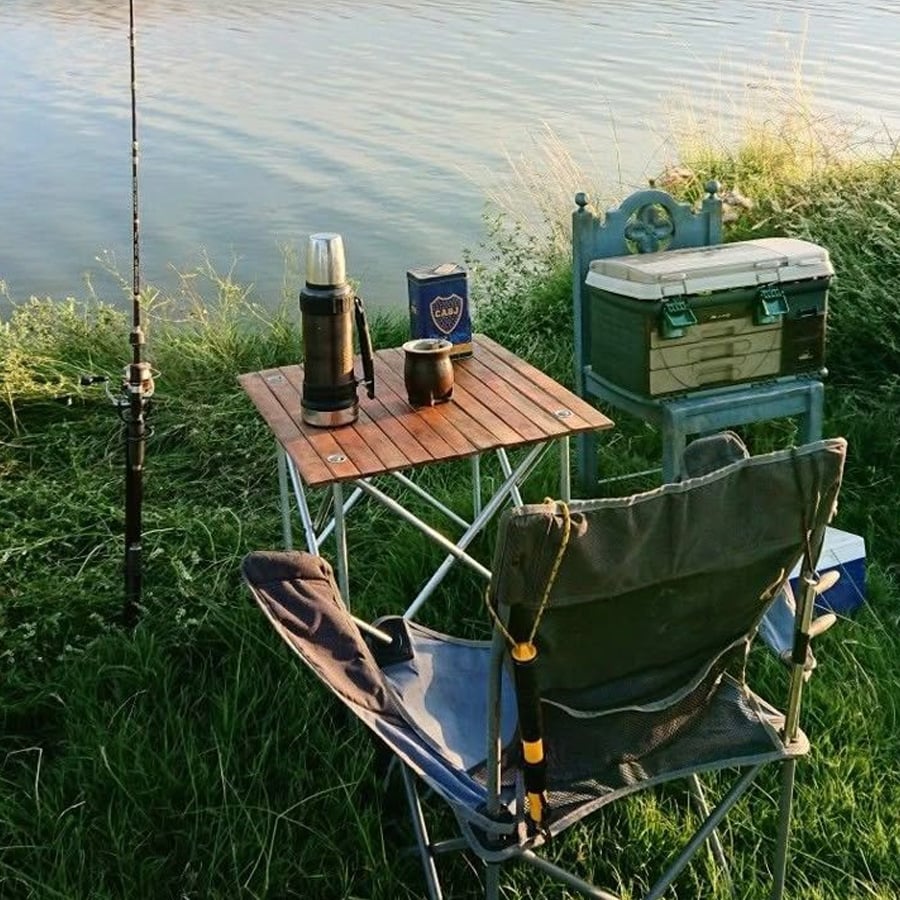 A fishing setup by a calm lake includes a folding chair, a small wooden table with a thermos and cup, a tackle box on a plastic chair, and a fishing rod propped up on the grassy shore.