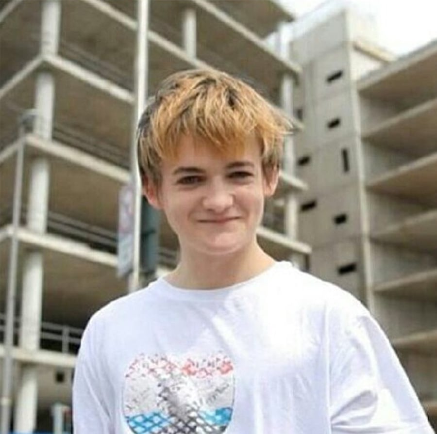 A young person with light brown hair, wearing a white T-shirt with a colorful graphic, stands outside in front of a multi-story concrete parking garage, smiling at the camera.