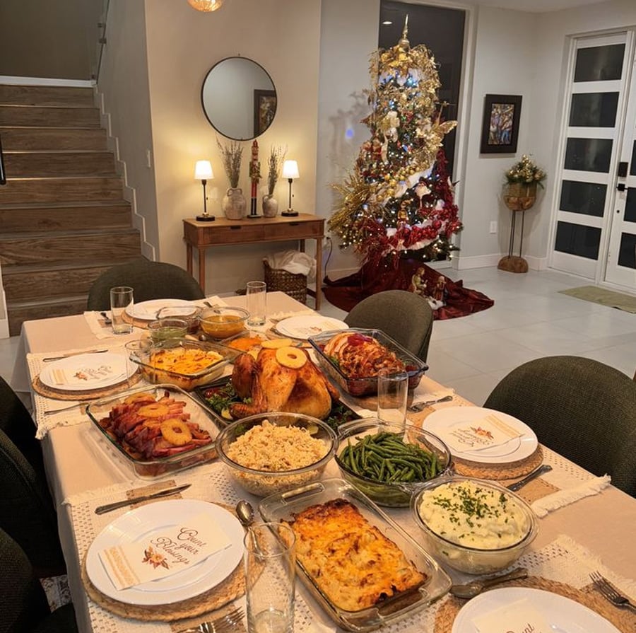 A dining table set for a festive meal features roasted meats, mashed potatoes, green beans, rice, and other dishes. In the background, a decorated Christmas tree and softly lit lamps enhance the cozy, holiday atmosphere.