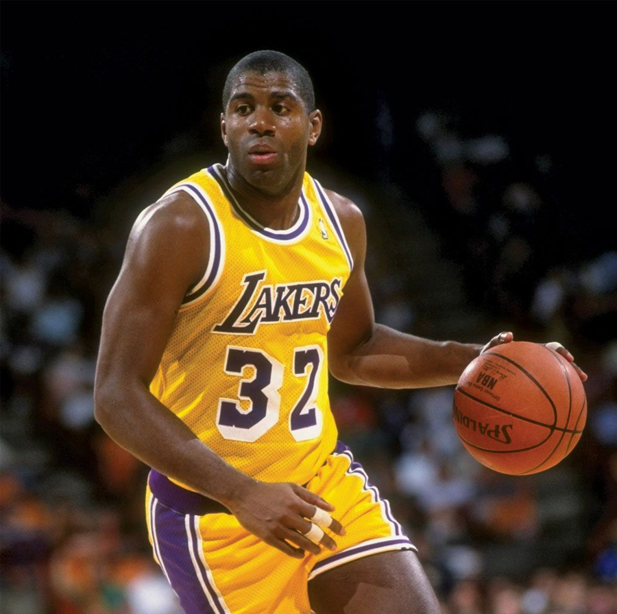 A basketball player wearing a yellow Lakers jersey with the number 32 dribbles a basketball on the court during a game. The background is blurred with the crowd in the stands.