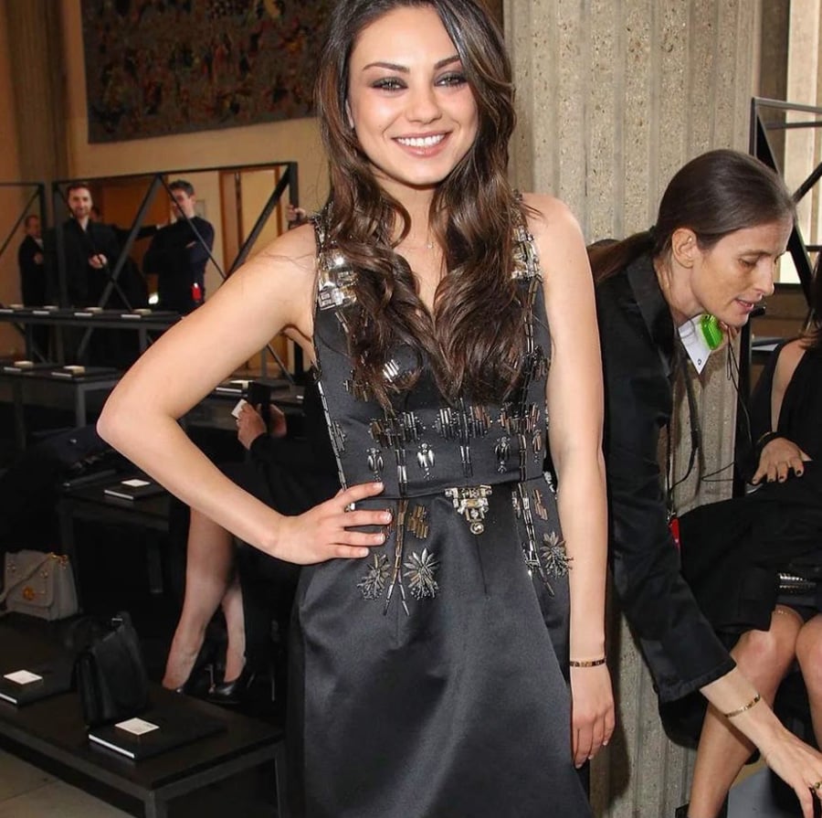 A woman with long brown hair wearing a sleeveless black dress with metallic embellishments stands smiling and posing indoors at an event, with people and empty seats in the background.