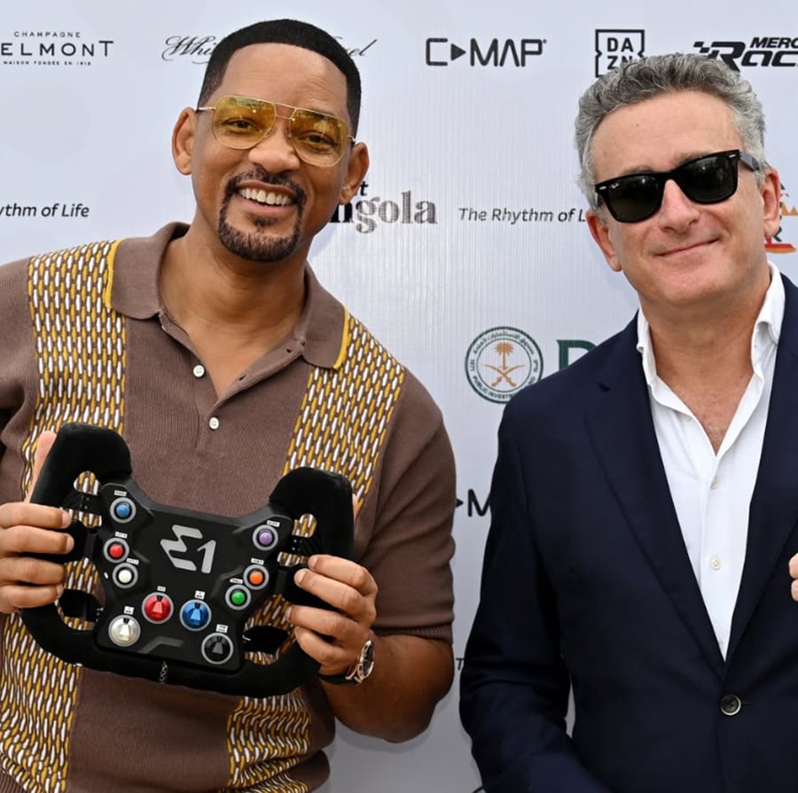 Will Smith, wearing a patterned shirt and sunglasses, smiles and holds a racing steering wheel labeled "E1" next to a man in a dark suit and sunglasses, standing in front of a backdrop with various sponsor logos.
