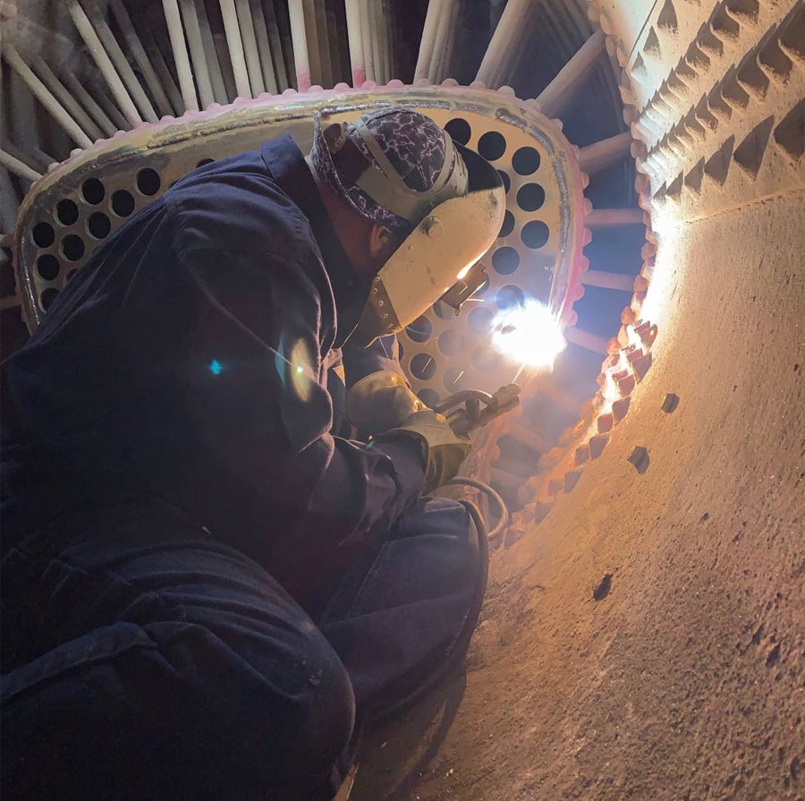 A person wearing protective gear welds inside a confined industrial space, illuminated by the bright light from the welding torch. Pipes and metal structures surround the worker.