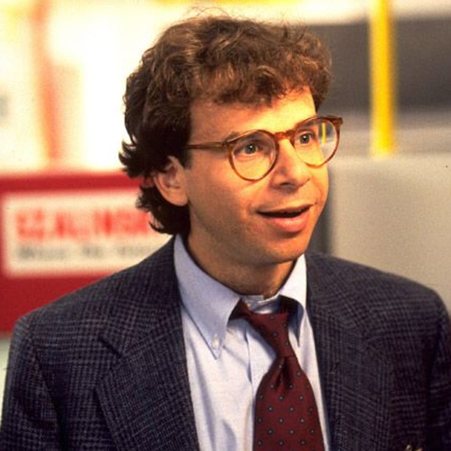A man with curly brown hair and glasses wears a blue checked suit, light blue shirt, and red polka dot tie, standing indoors and smiling slightly.