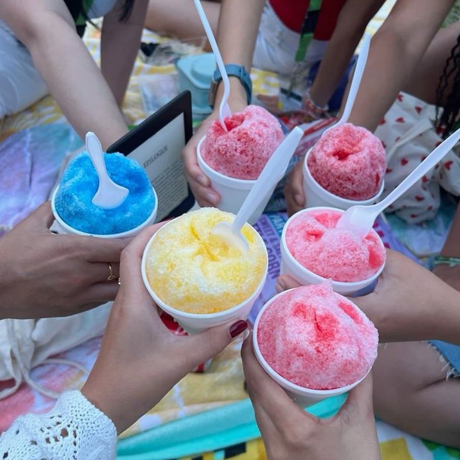 A group of people holding colorful shaved ice cups—blue, yellow, and pink—close together as if making a toast. They are outdoors, sitting on a blanket, with spoons in each cup.