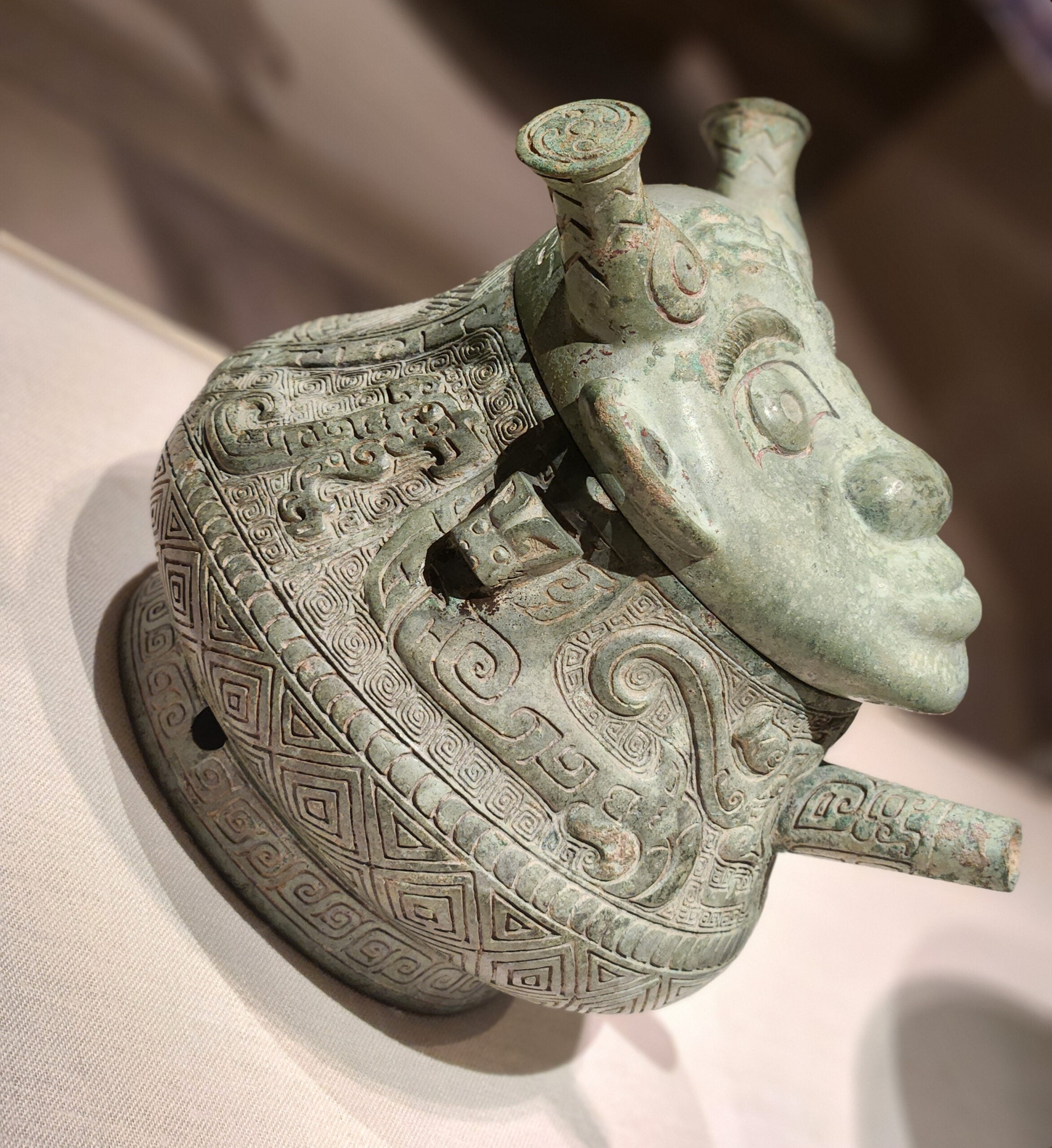 A bronze vessel with intricate geometric and animal patterns, featuring a sculpted human-like face and horns, displayed on a light-colored surface in a museum-like setting.