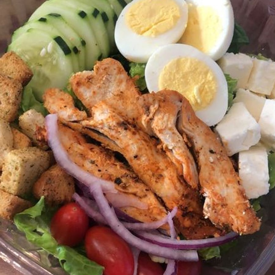 A salad with grilled chicken strips, sliced hard-boiled egg, cucumber, croutons, feta cheese cubes, grape tomatoes, red onion slices, and lettuce in a clear container.