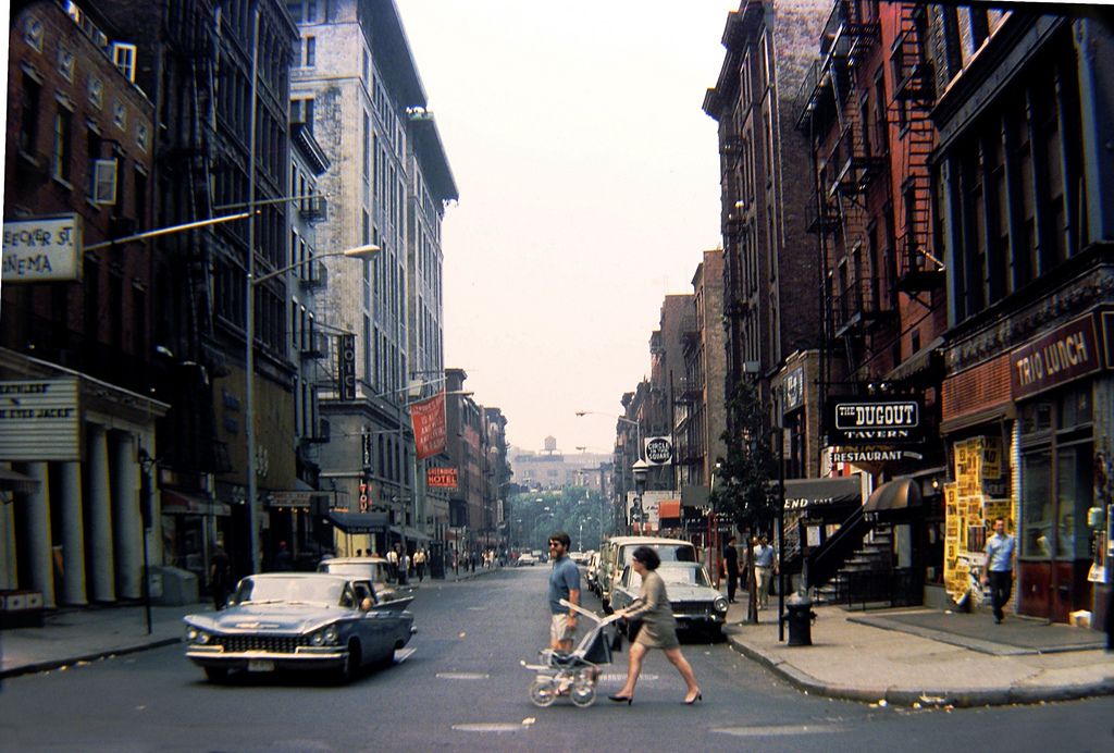 A city street scene from the mid-20th century shows people crossing the road with a baby stroller, vintage cars parked and driving, and storefronts with colorful signs lining both sides of the street.