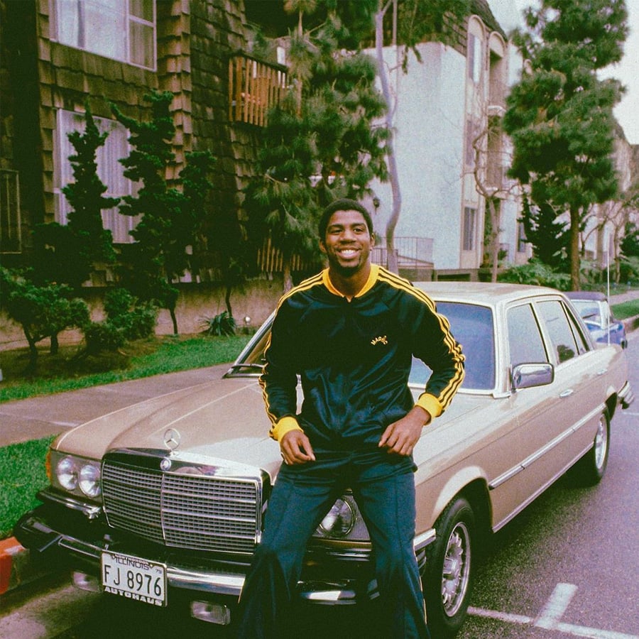 A man wearing a dark tracksuit with yellow stripes sits smiling on the hood of a beige Mercedes-Benz parked on a residential street lined with trees and apartment buildings.