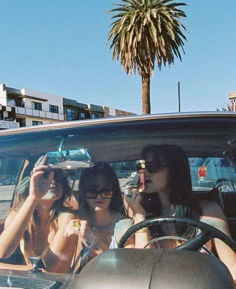 Three young women sit in a car under a clear blue sky with a tall palm tree visible outside. Two wear sunglasses, and one applies lipstick while looking in the rearview mirror. Apartment buildings are in the background.