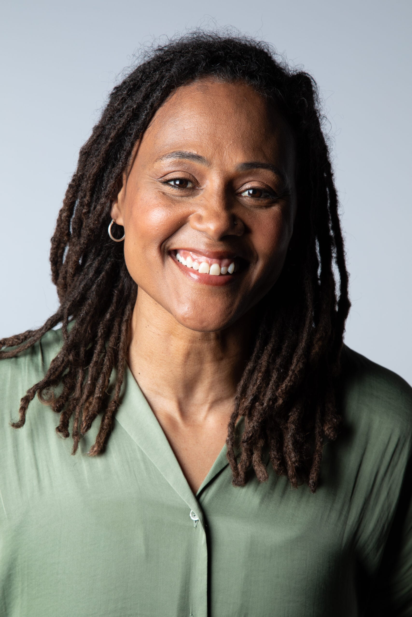 A woman with long dreadlocks, wearing a light green button-up shirt, smiles at the camera against a plain light gray background.