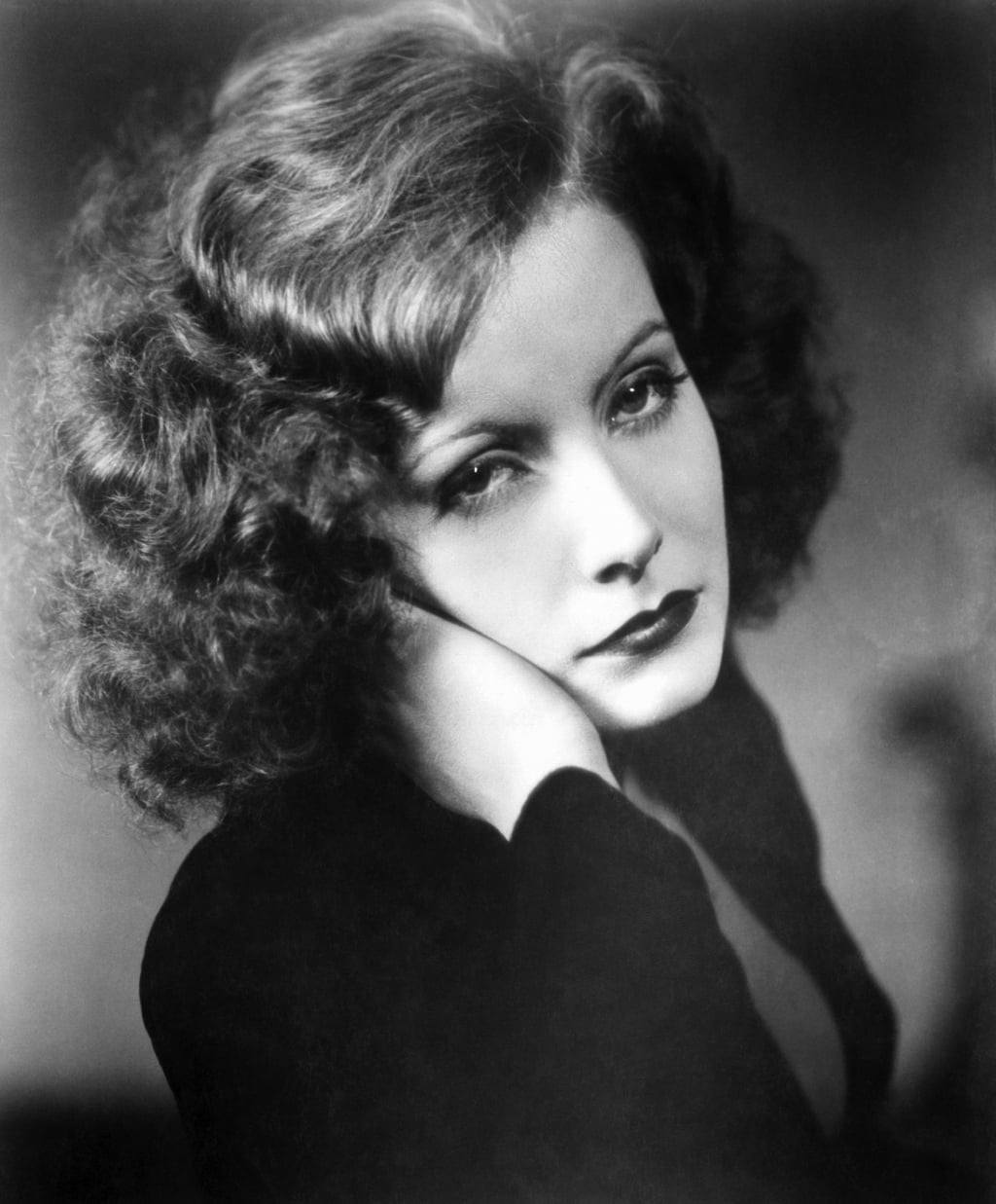 Black-and-white portrait of a woman with wavy hair resting her head on her hand, gazing thoughtfully at the camera with an expression of melancholy or contemplation.