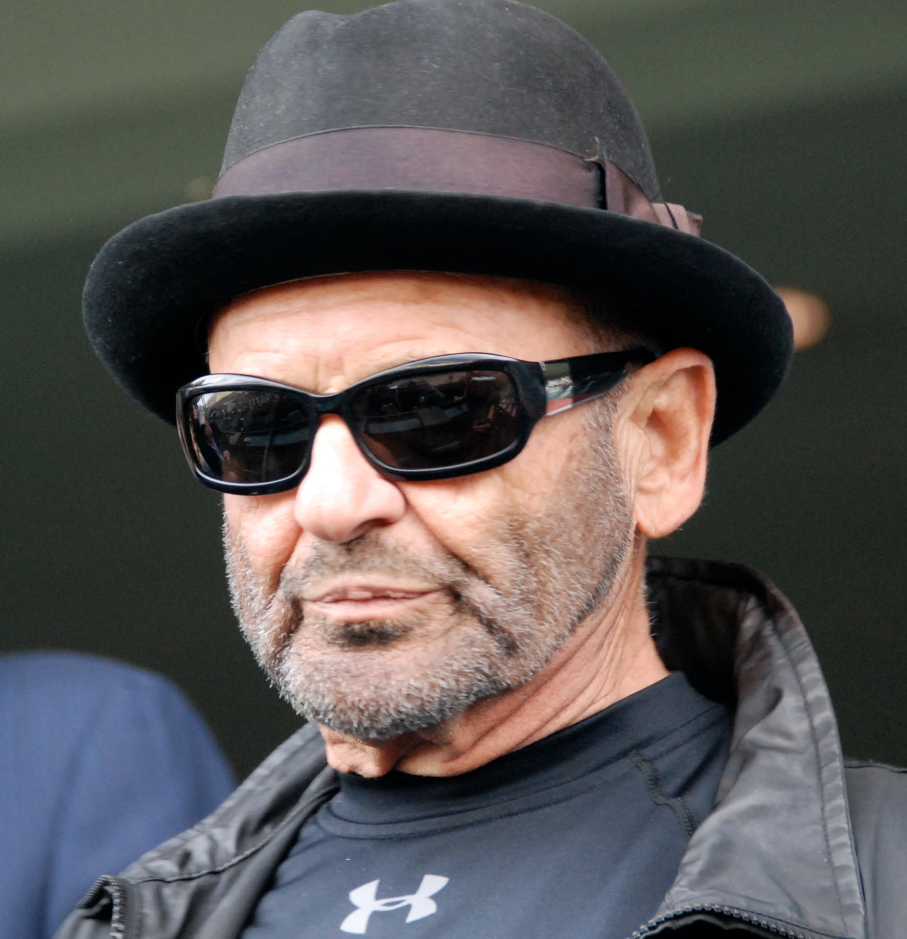 A man with a trimmed beard wears a black fedora, dark sunglasses, a black Under Armour shirt, and a black leather jacket. The background is blurred.